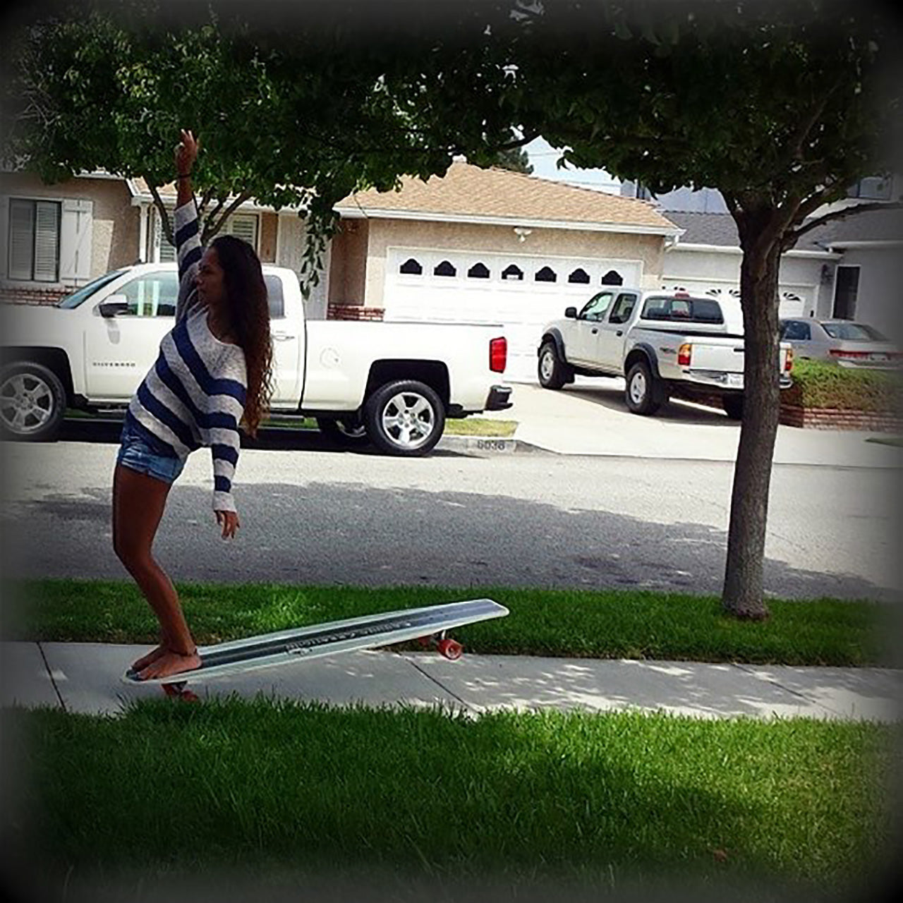 Woman riding a Kahuna Creations longboard on a sidewalk in a neighborhood with grass and trees