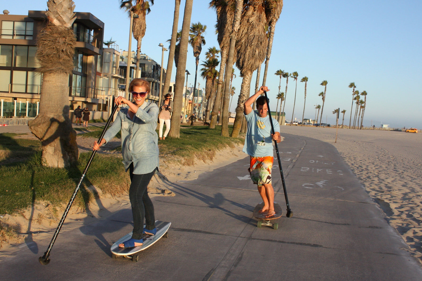 Man and woman riding Kahuna Creations longboards down a beach path lined by palm trees.