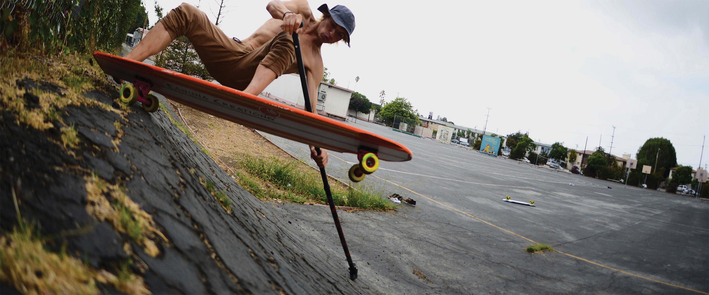 Man riding Kahuna Creations Bombora longboard skateboard with 180mm trucks, 70mm wheels and ABEC7 bearings. Using a land paddle big stick skate pole.