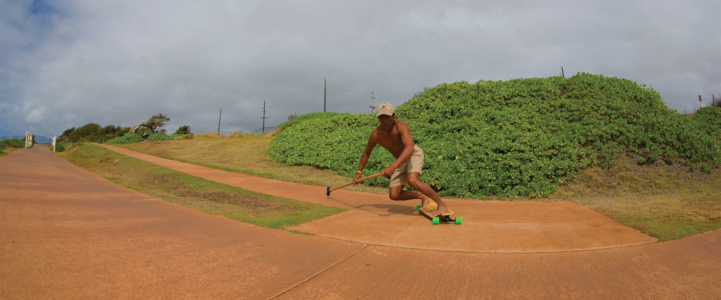 Man riding Kahuna Creations drop deck longboard skateboard, using a land paddle big stick skate pole in Hawaii. Using 180mm trucks, 69mm wheels and ABEC7 bearings.